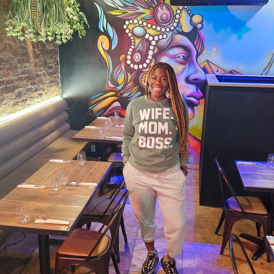 Woman in a green sweatshirt standing in a restaurant with colorful mural and wooden tables.