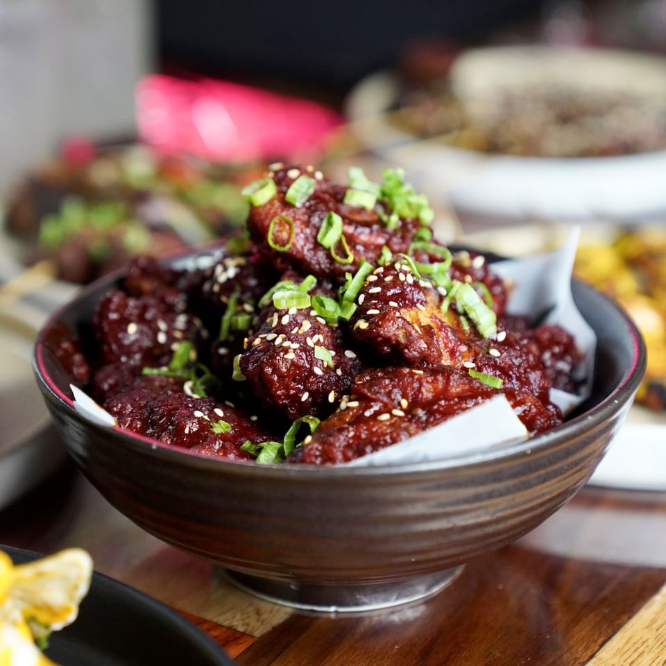 A dark bowl of saucy chicken wings, garnished with sesame seeds and chopped green onions.
