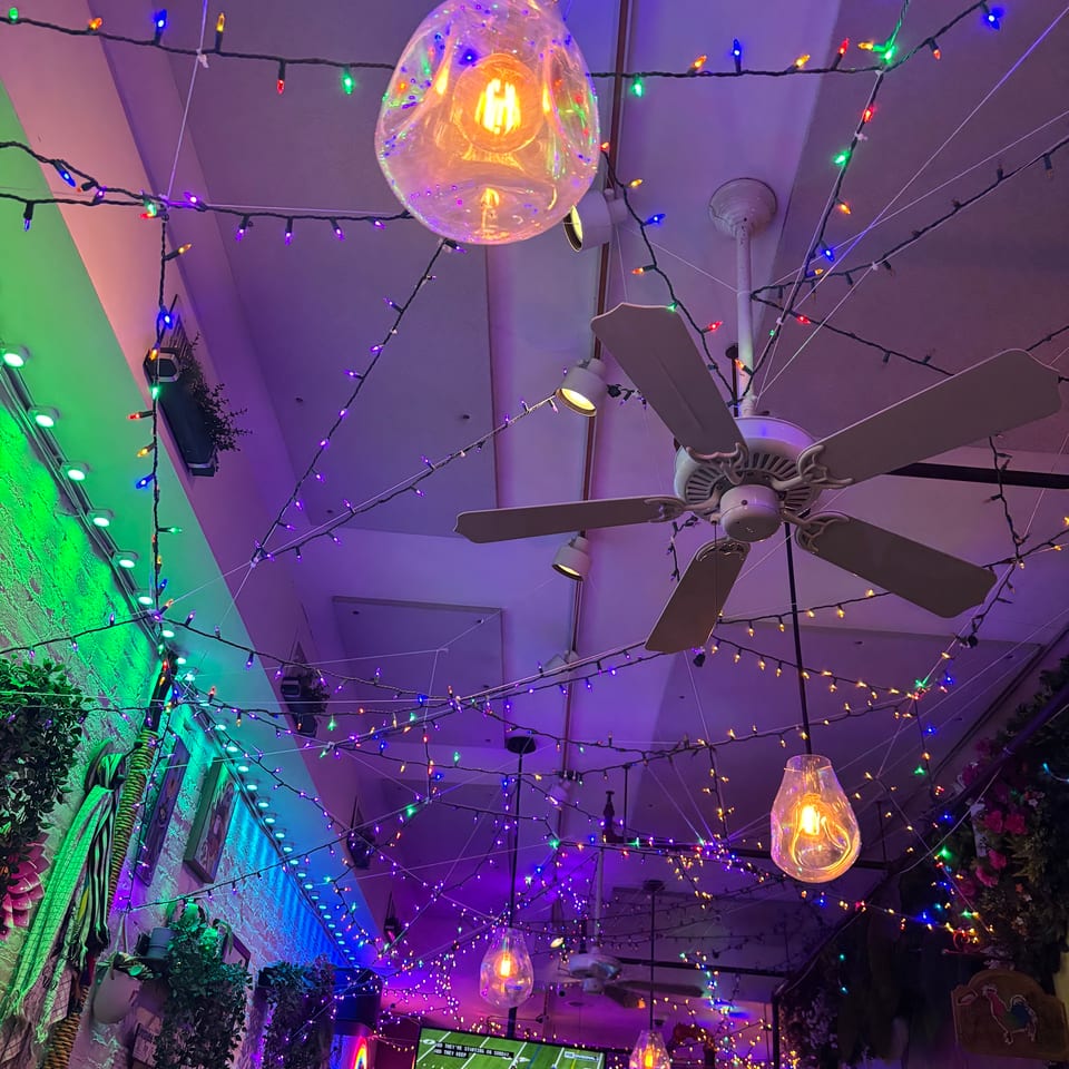 A restaurant ceiling adorned with colorful string lights, hanging bulbs, and ceiling fans, with a TV showing football.