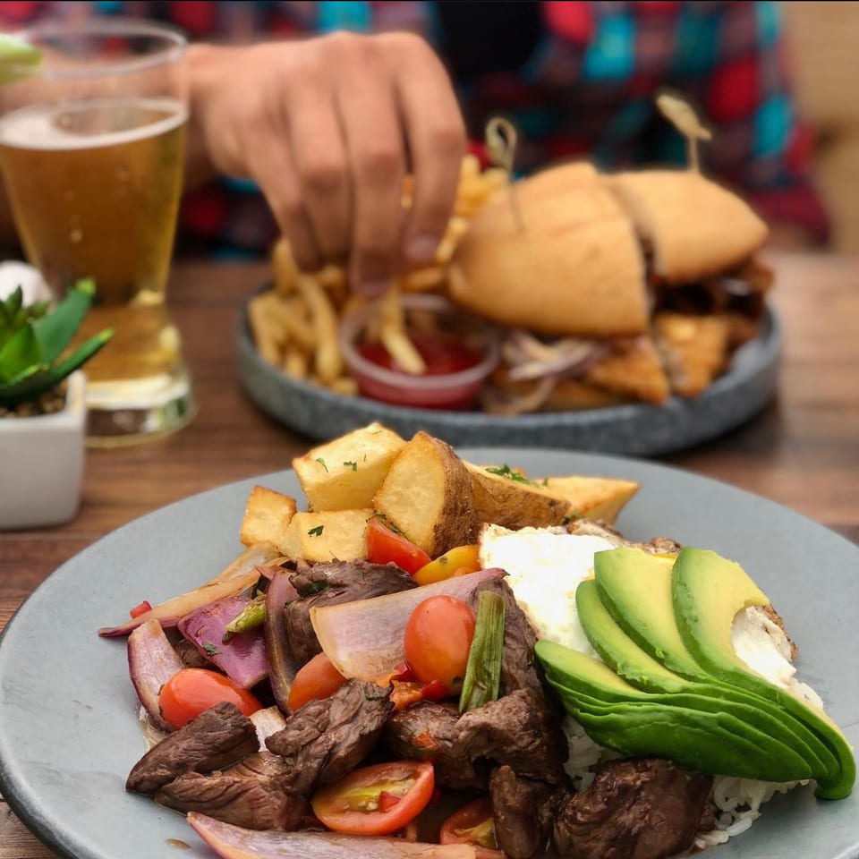Peruvian Lomo Saltado with avocado and fried egg, with a person eating burgers and fries in the background.