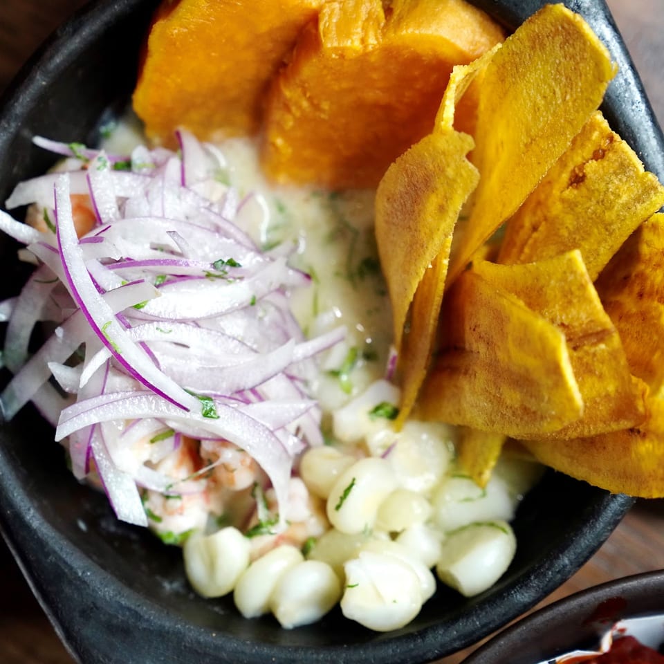 A close-up of ceviche with shrimp, red onions, corn, and plantain chips in a dark bowl.