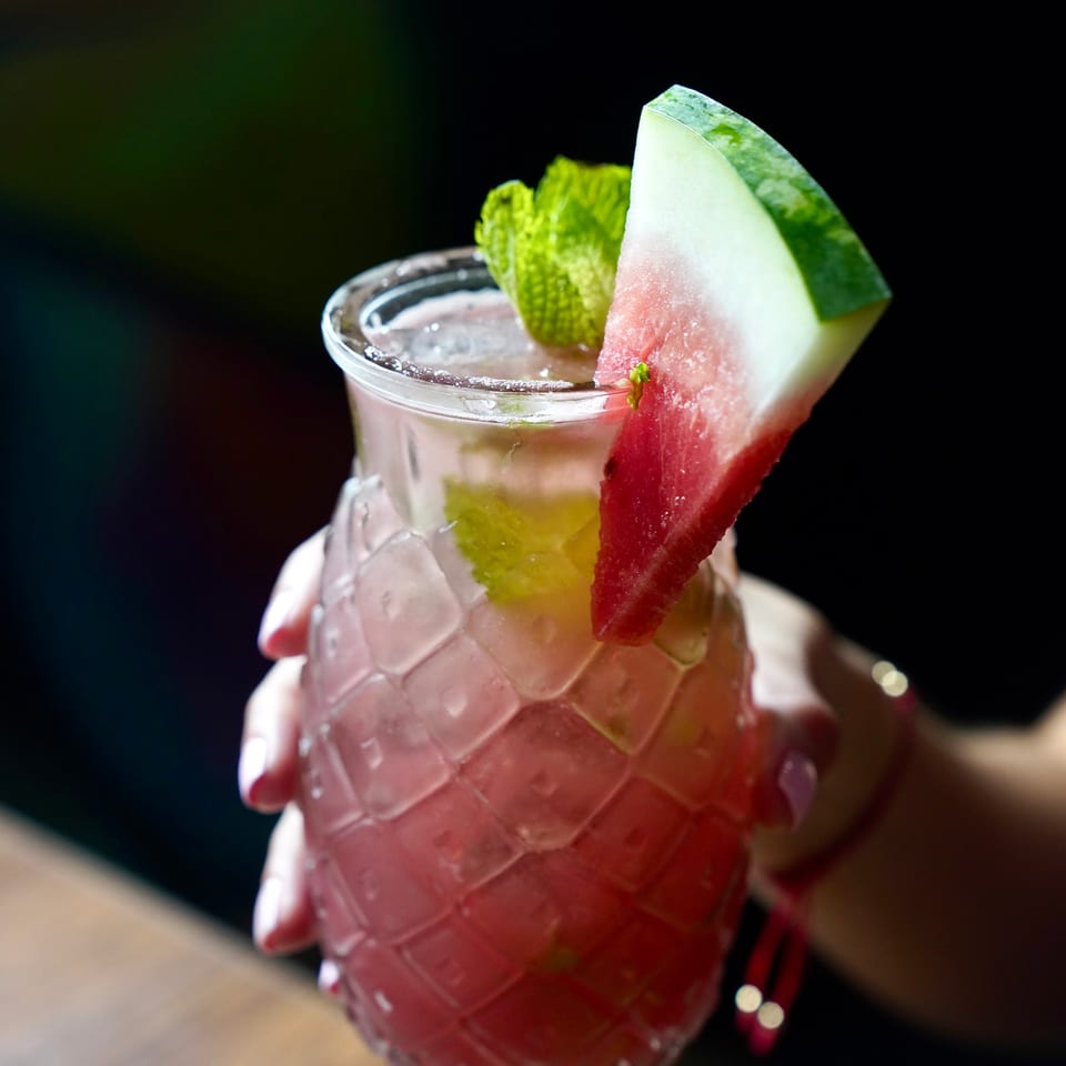 A hand holds a pink watermelon cocktail garnished with mint and a watermelon slice.