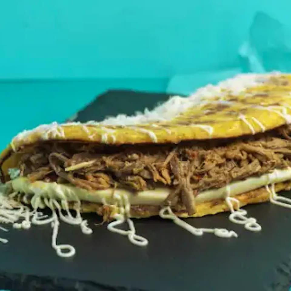 Arepa with shredded beef, cheese, and white sauce on a black plate with a blue background.