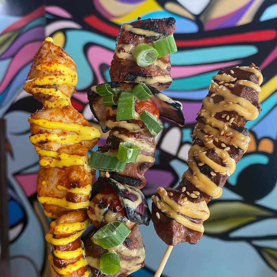 Three skewers of grilled meat and vegetables with colorful sauces and garnishes against a vibrant background.