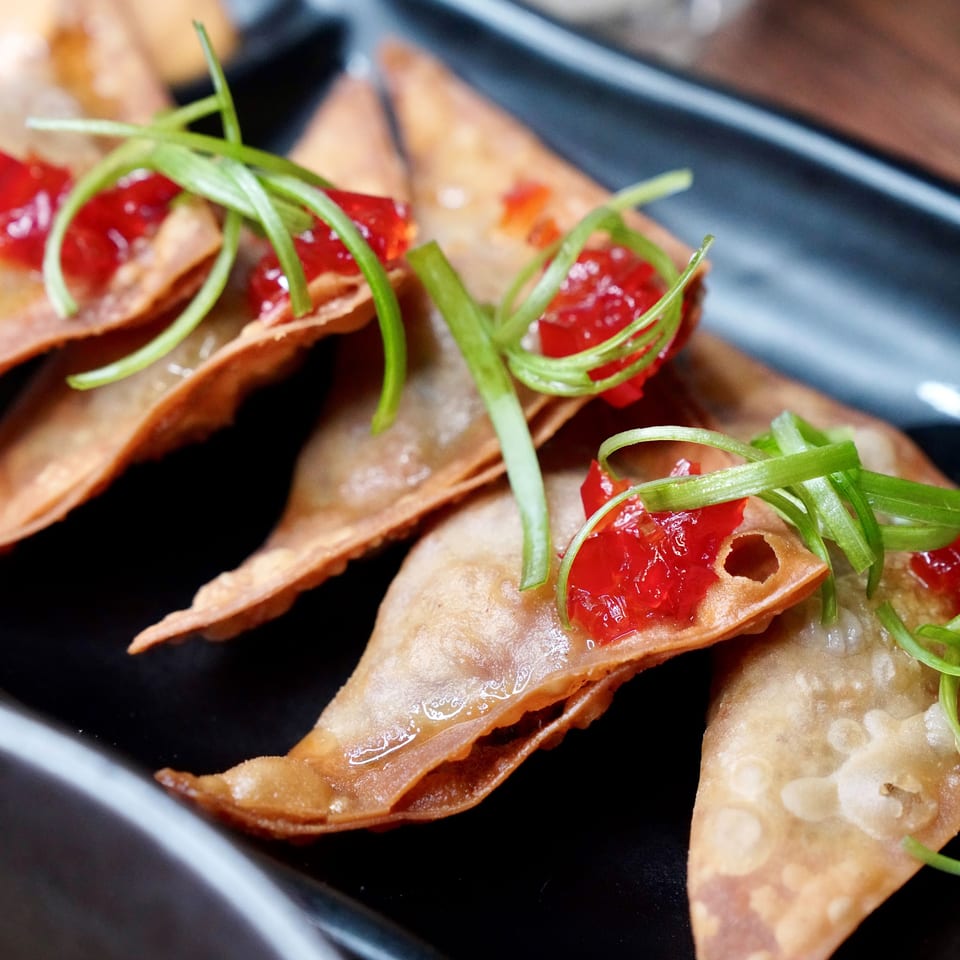 Crispy fried wontons topped with red jelly and green onions on a black rectangular plate.