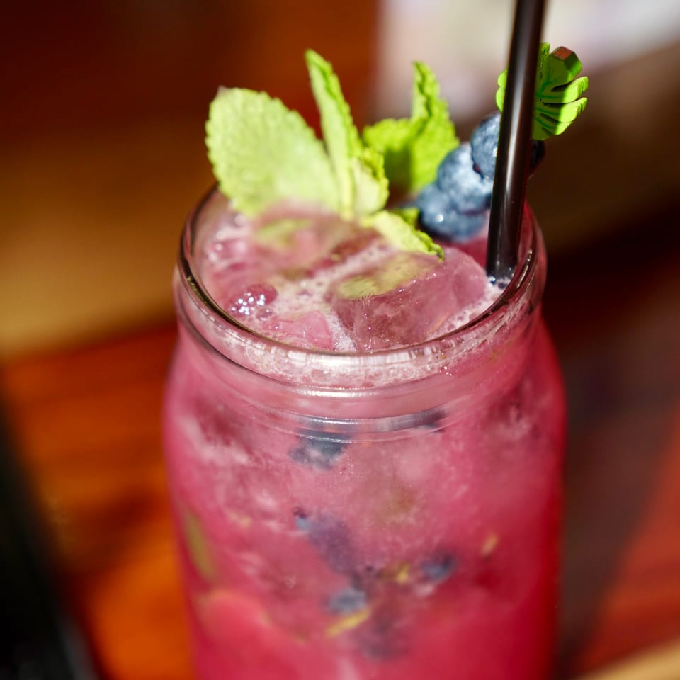 A vibrant pink cocktail with ice, blueberries, and mint leaves, served in a glass jar.