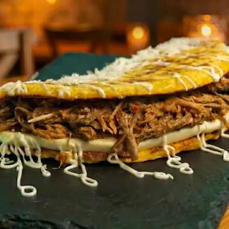 Arepa filled with shredded beef, cheese, and a drizzle of white sauce on a dark slate.